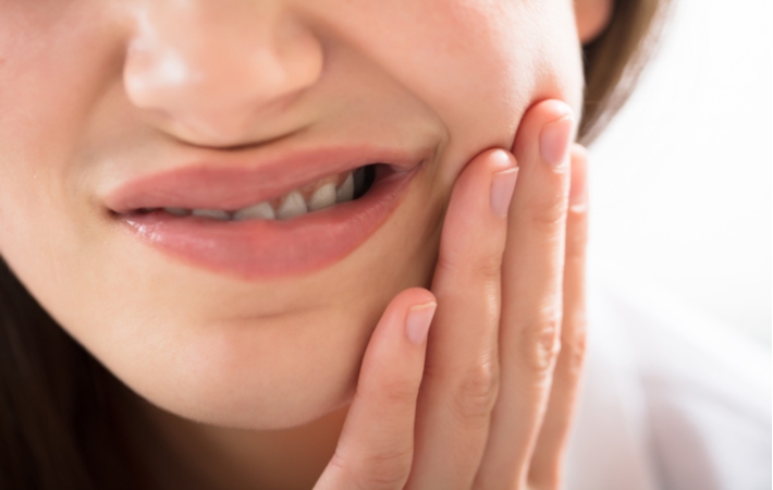 A close up of a woman holding her teeth in pain as she is dealing with the discomfort of tooth sensitivity