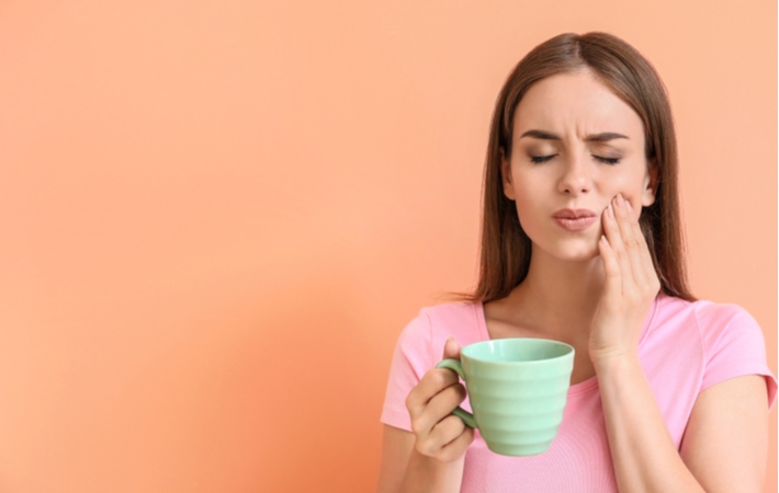 A woman in front of an orange background with a pink t-shirt, drinking coffee out of a green mug holding her cheek in pain from her sensitive teeth
