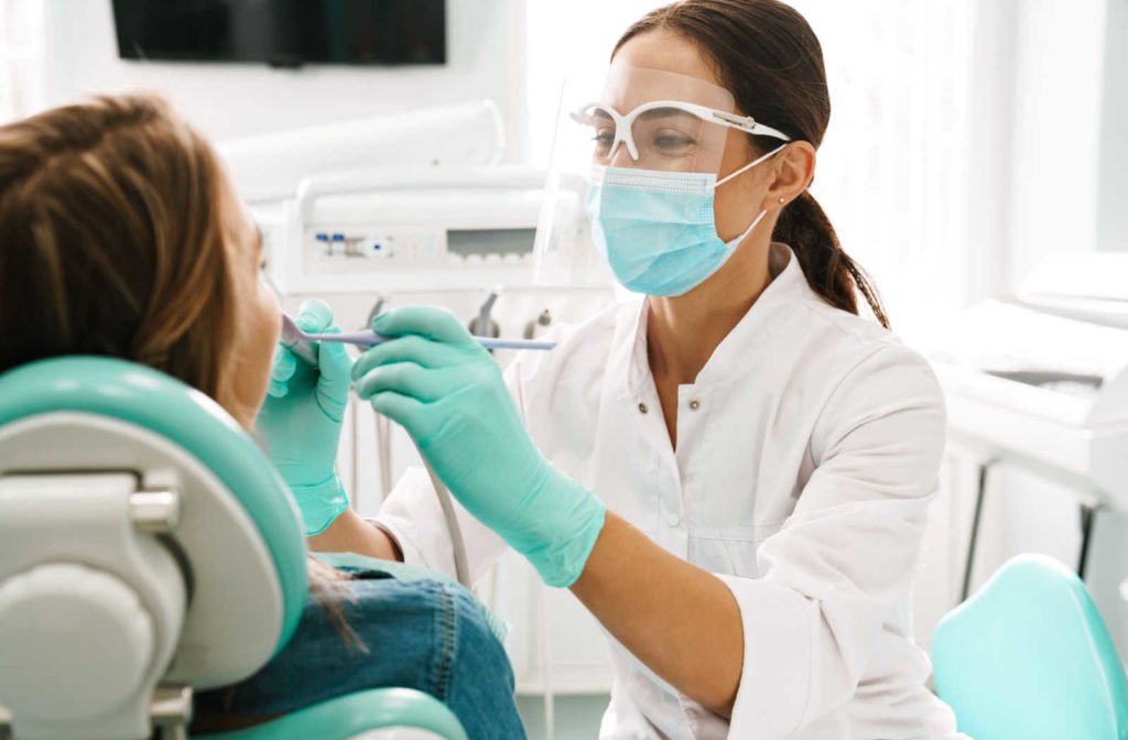 A female dentist wearing a mask and face shield holds dental tools while working on a patient.