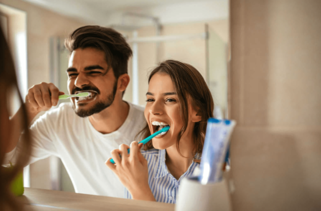 A couple smiling in the mirror of their bathroom while they brush their teeth to maintain good oral hygiene between dental cleanings