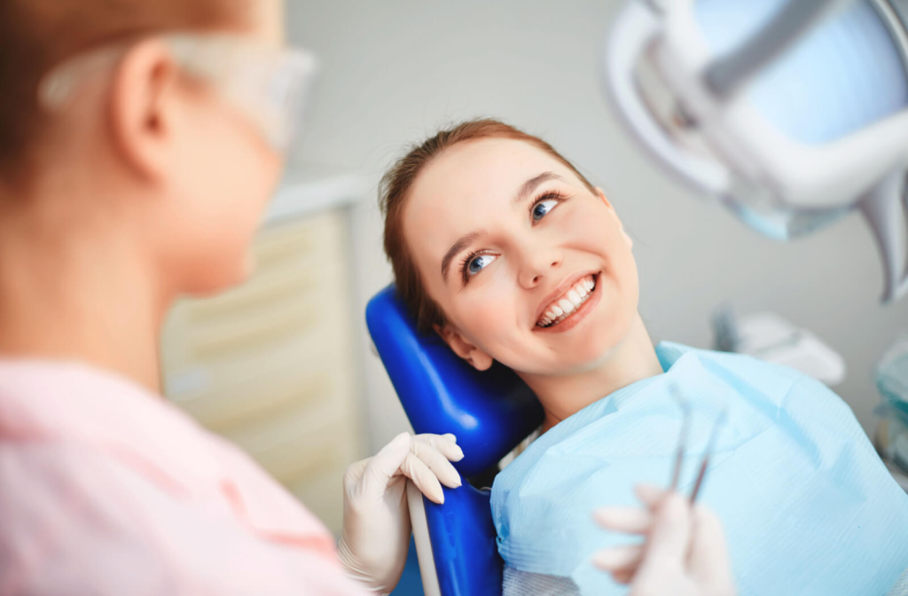 A young child sitting in a dentist's chair smiling while her dentist examines her teeth.