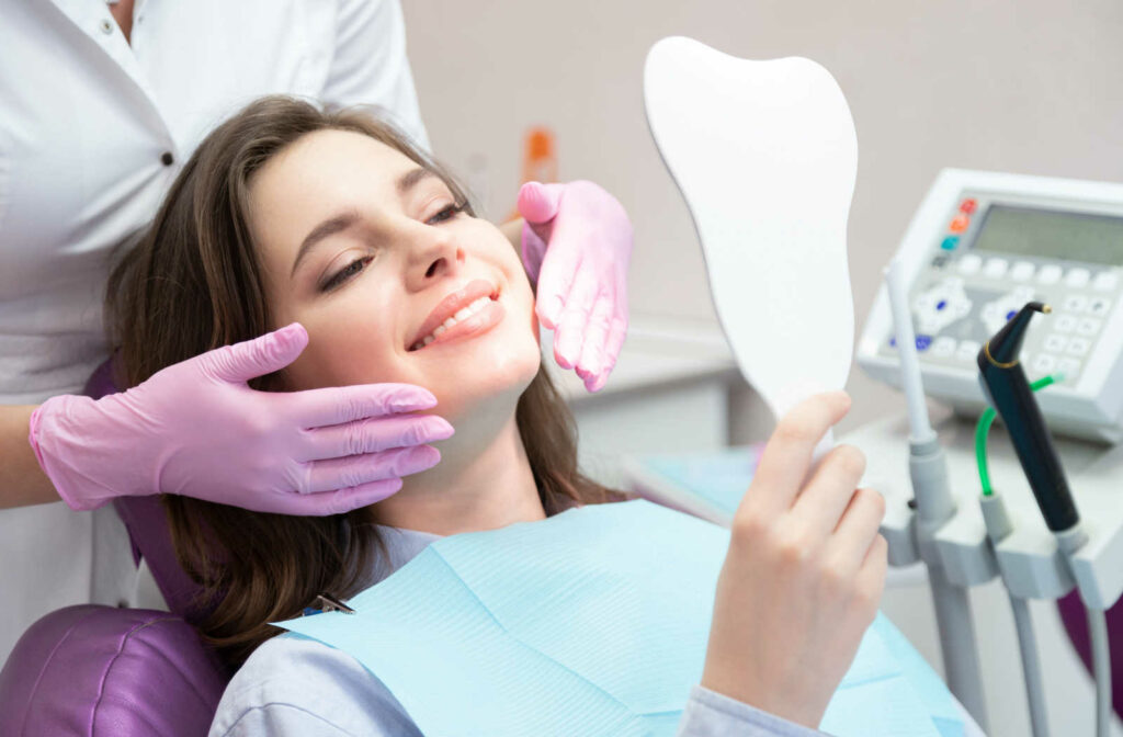 A woman sitting in a dentist’s chair and smiling into a handheld mirror while her dentist gently holds the sides of her face