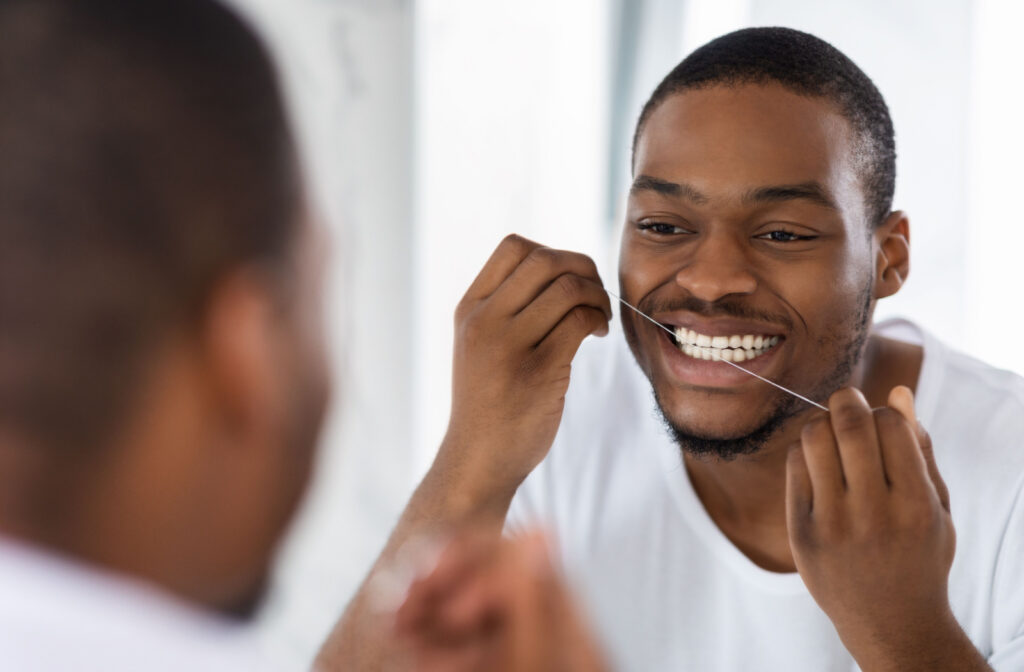 A young man in a white shirt flossing his teeth in front of a mirror.