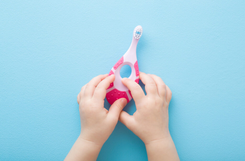 A close-up of a baby's hands holding a pink toothbrush with a wide handle made for babies.