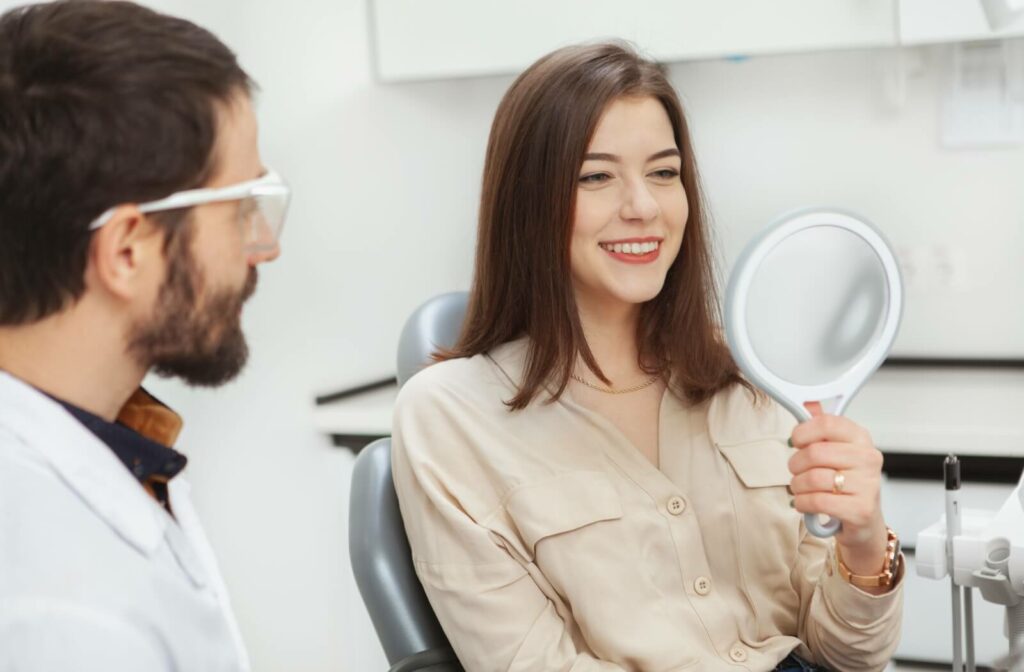 An adult smiling in a handheld mirror after having dental implants installed while their dentist explains what to expect during recovery.