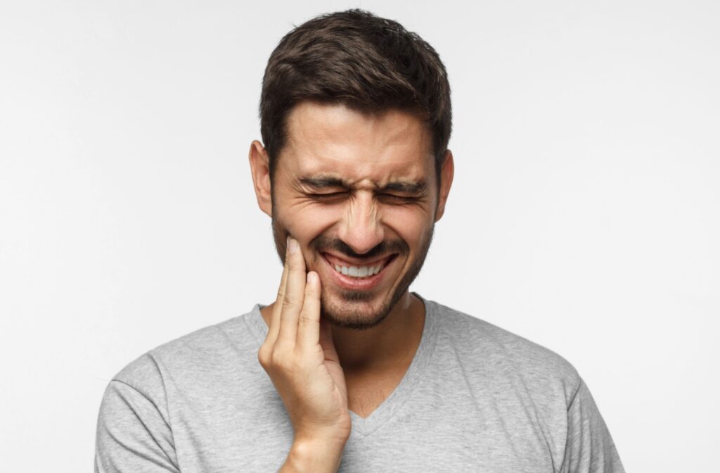 Man in grey shirt holding his hand to the side of his face with a wincing expression.