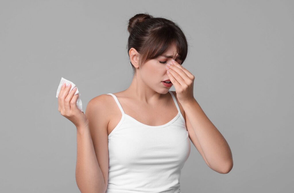 Woman in white tank top holding bridge of her nose in pain with a tissue in her other hand.